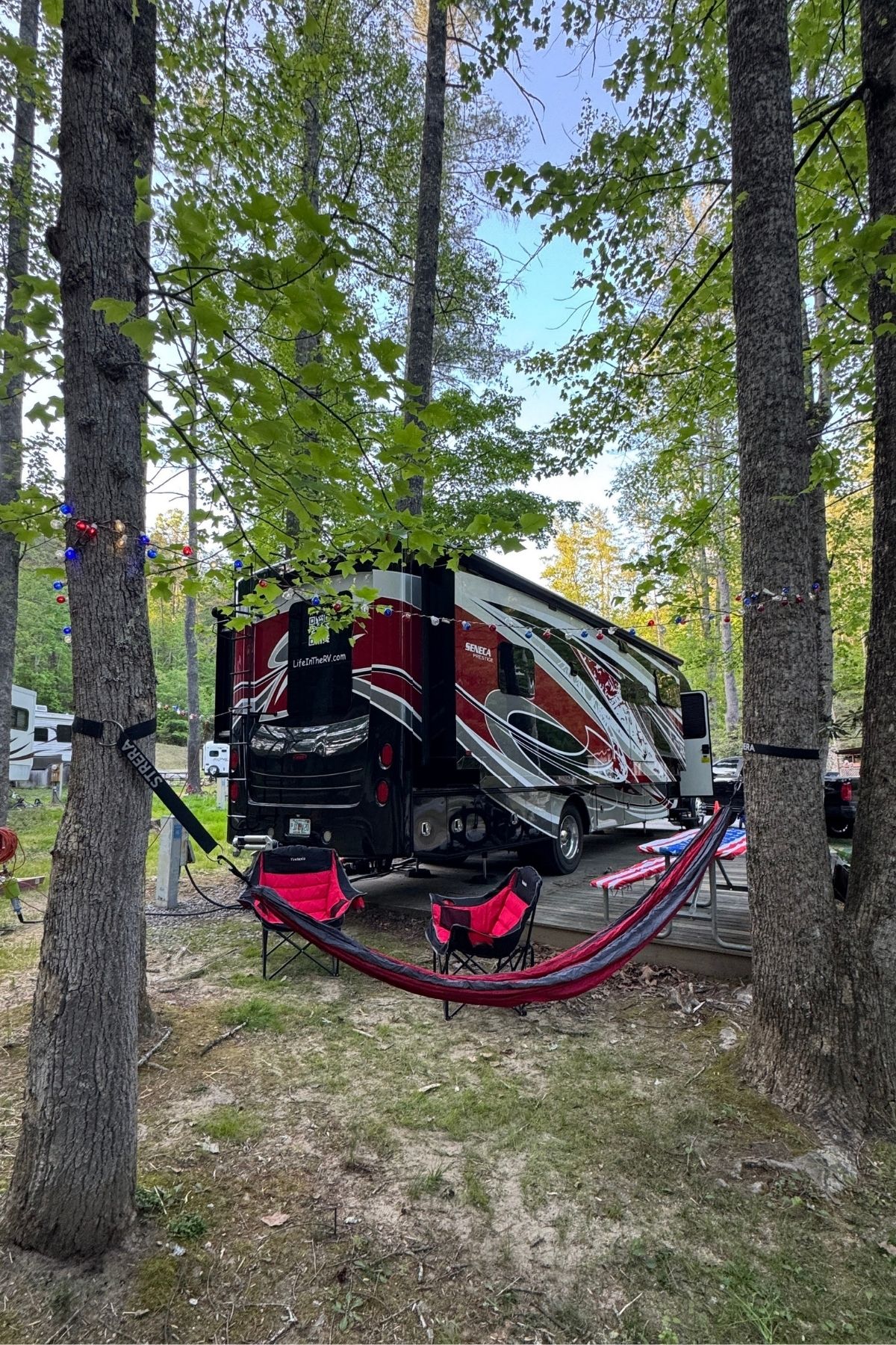 A large red and white RV is parked at a wooded Green Mountain Thousand Trails campsite, with two red hammocks strung between trees and two red chairs set up nearby.