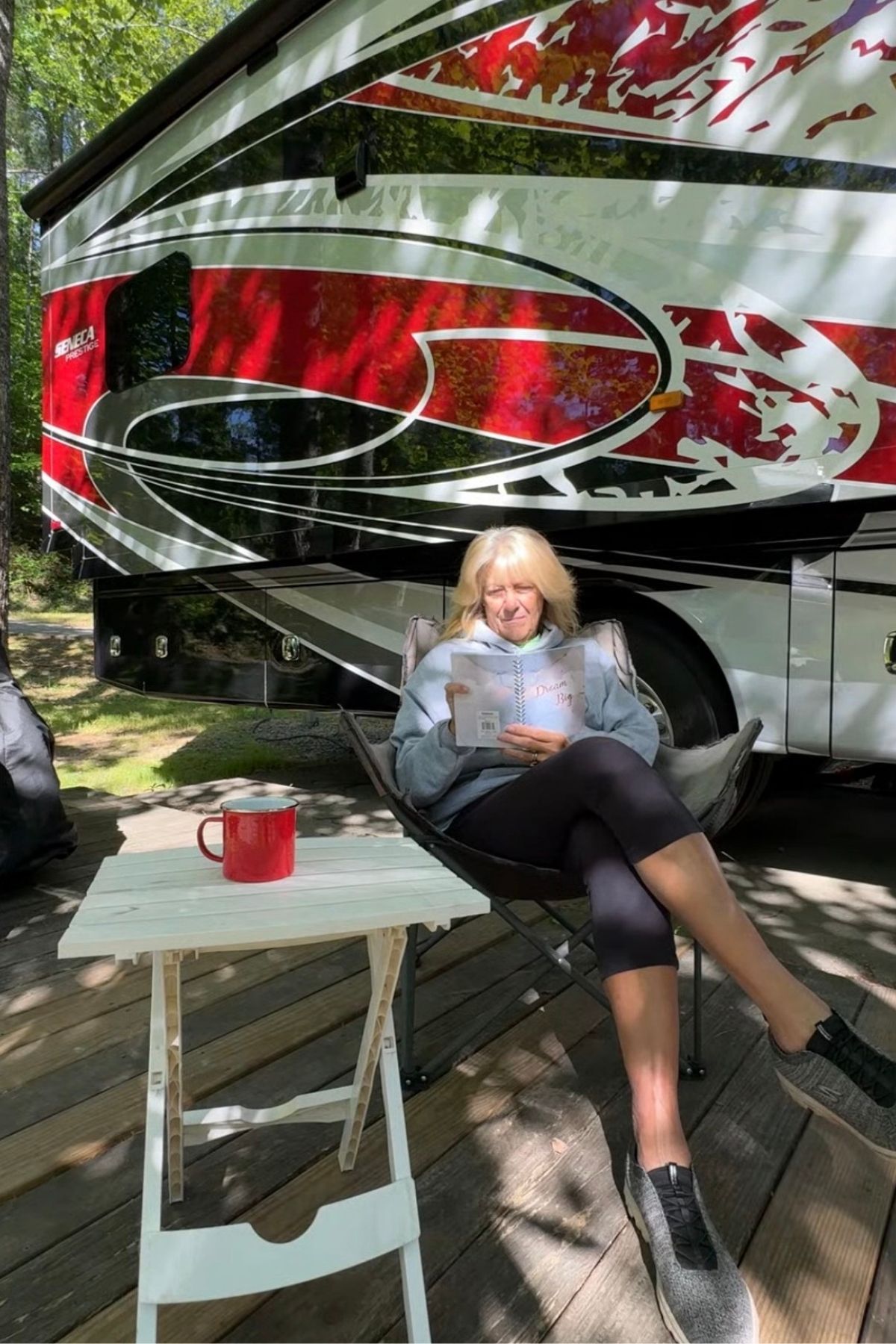 A woman sits in a camping chair reading a book next to a table with a red mug, outside a large RV parked on a wooden deck at Green Mountain Thousand Trails in a peaceful wooded area.