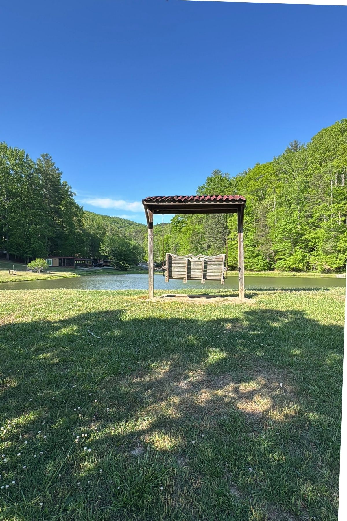 A wooden swing with a roof sits on a grassy area by the lake at Green Mountain Thousand Trails, surrounded by lush green trees under a clear blue sky.
