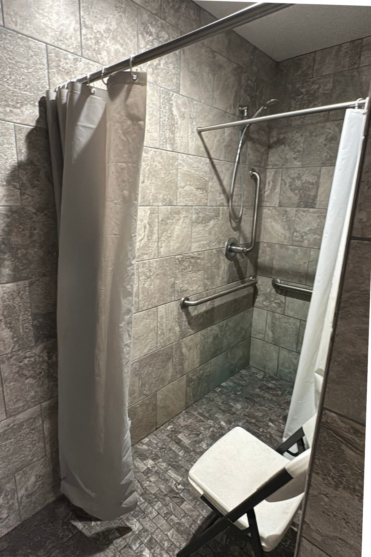 Accessible shower with gray tiled walls, grab bars, a handheld showerhead, shower curtain, and a white folding chair—perfect for guests at Green Mountain Thousand Trails seeking comfort and convenience.
