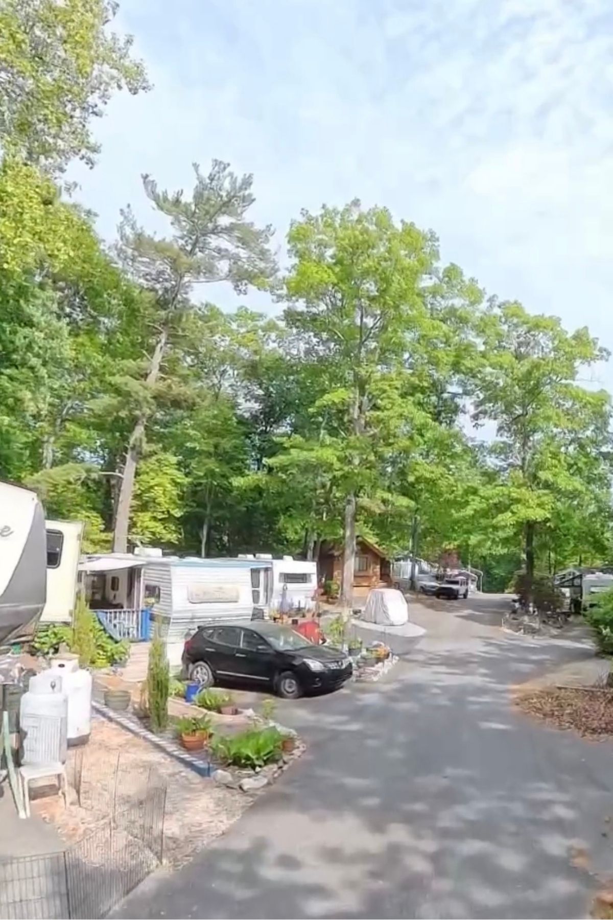 A paved road winds through Green Mountain Thousand Trails campground, passing parked RVs, a black car, potted plants, and tall green trees beneath a partly cloudy sky.