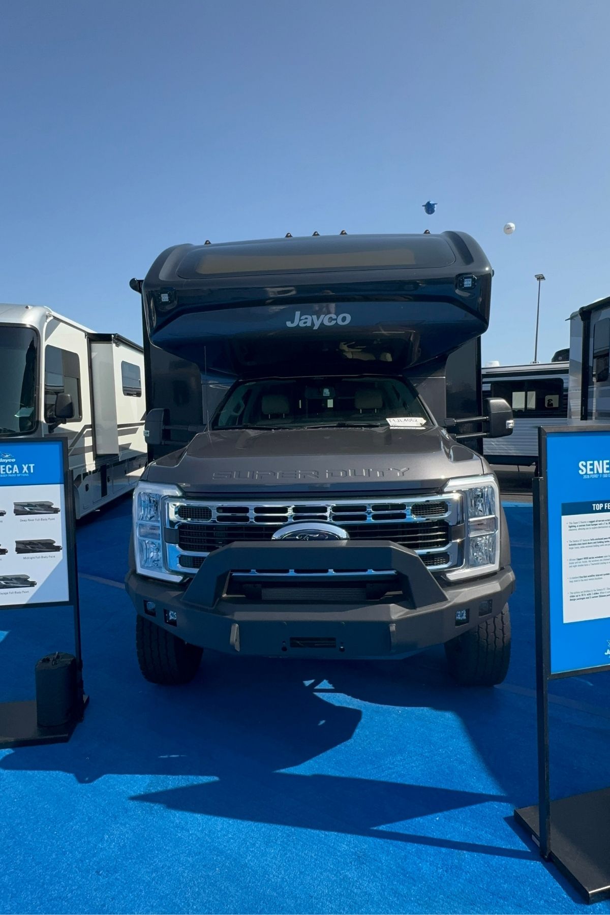 A Jayco Seneca XT, one of the top Jayco Class C Motorhomes, is displayed on a blue carpet outdoors, flanked by informational signs and surrounded by other RVs in the background.