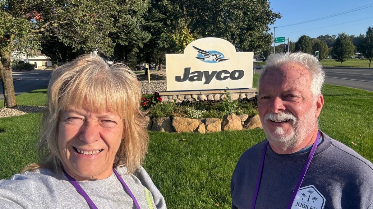 Two smiling older adults stand outside in front of a "Jayco" sign, with green grass, trees, and a road in the background—ready to embark on adventures in their Jayco Class C Motorhomes.