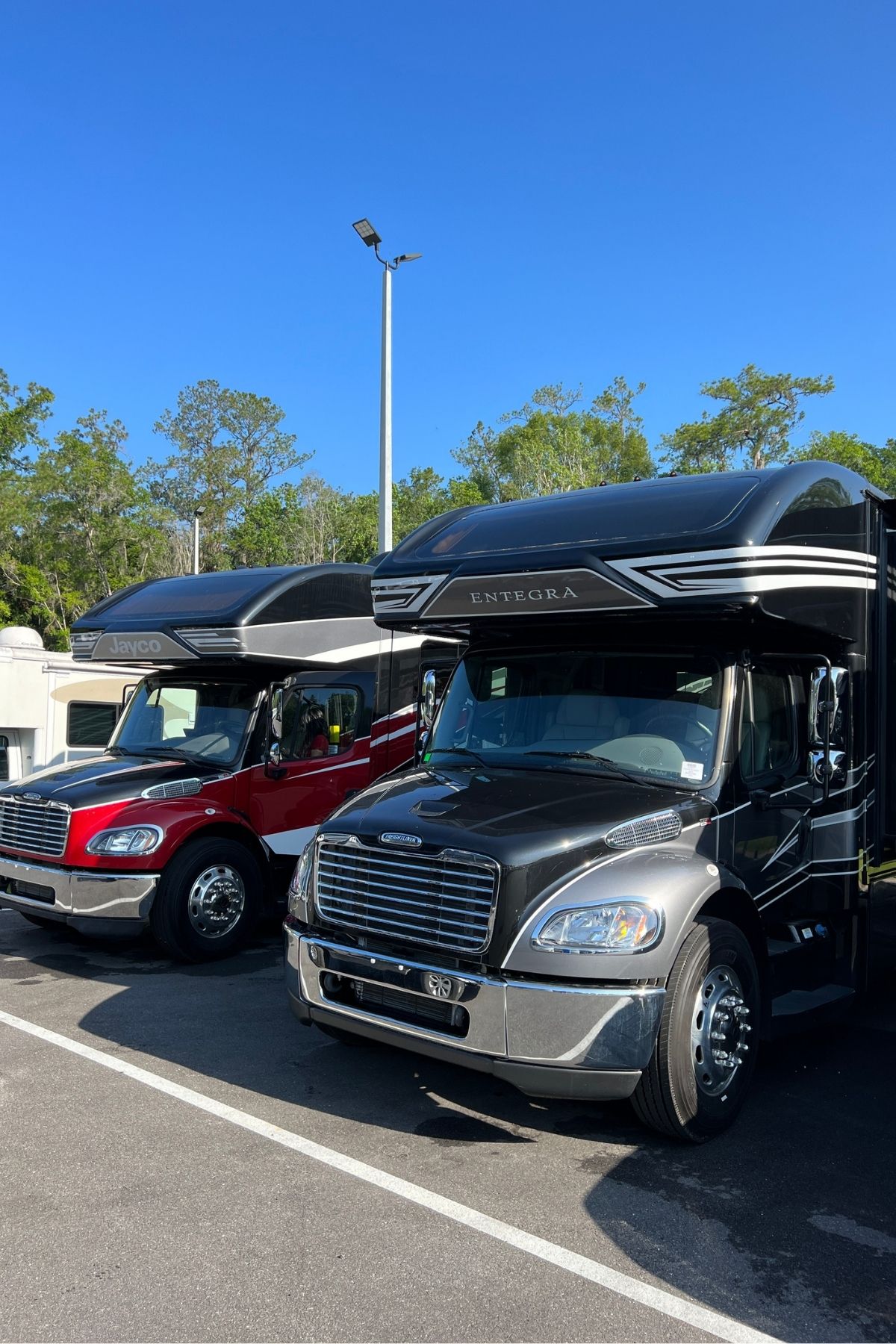 Two large RV motorhomes, including stylish Jayco Class C Motorhomes—one black and one red—are parked side by side in a parking lot under a clear blue sky with trees in the background.