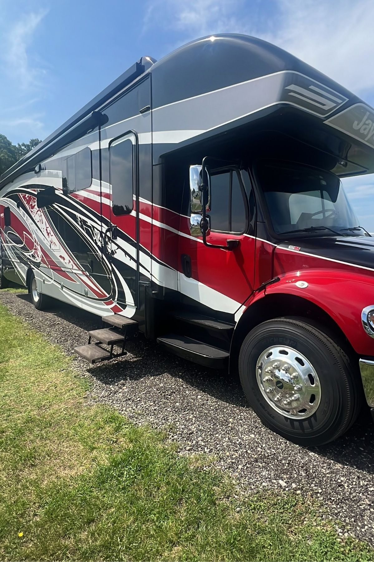 Large red, black, and white Jayco Class C Motorhome parked on a gravel surface beside grass under a blue sky.
