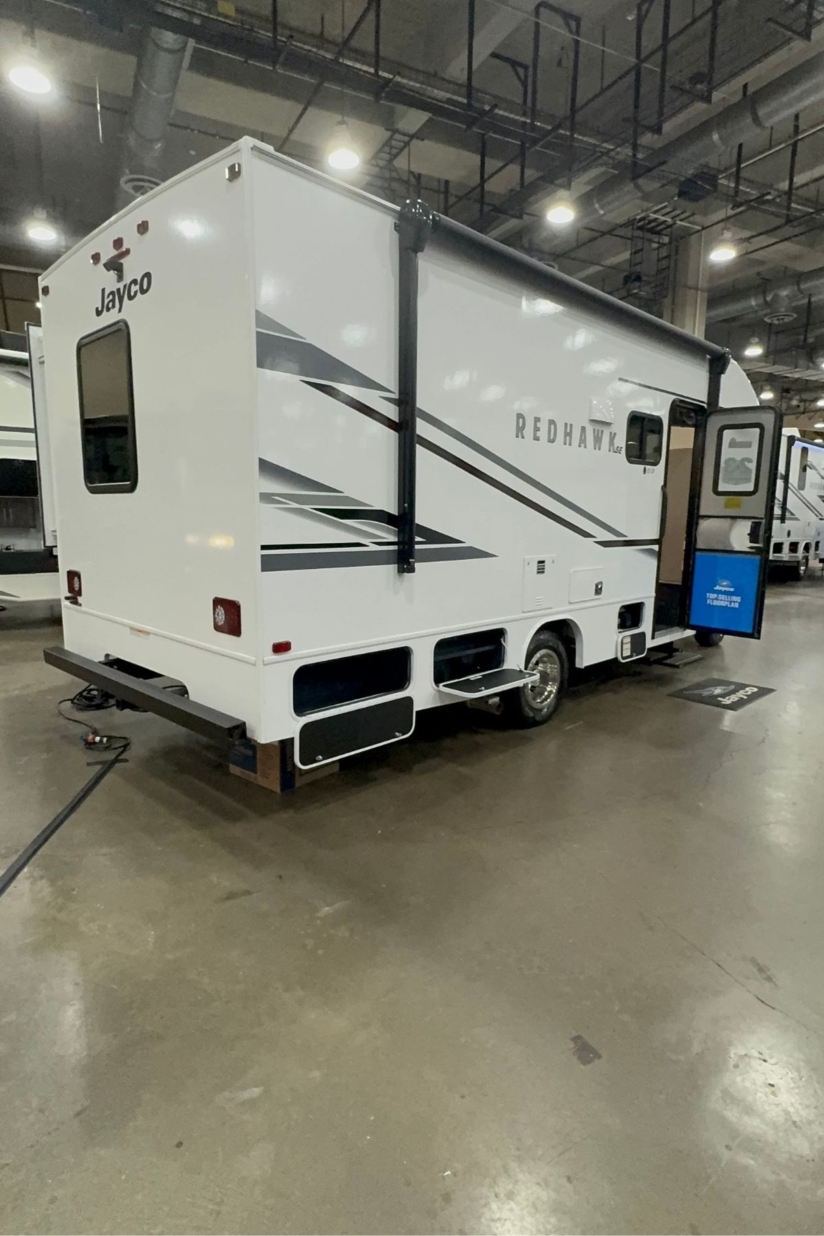 A white Jayco Redhawk, part of the Jayco Class C Motorhomes lineup, is parked indoors on a concrete floor with its entry door open and a power cable connected at the rear.