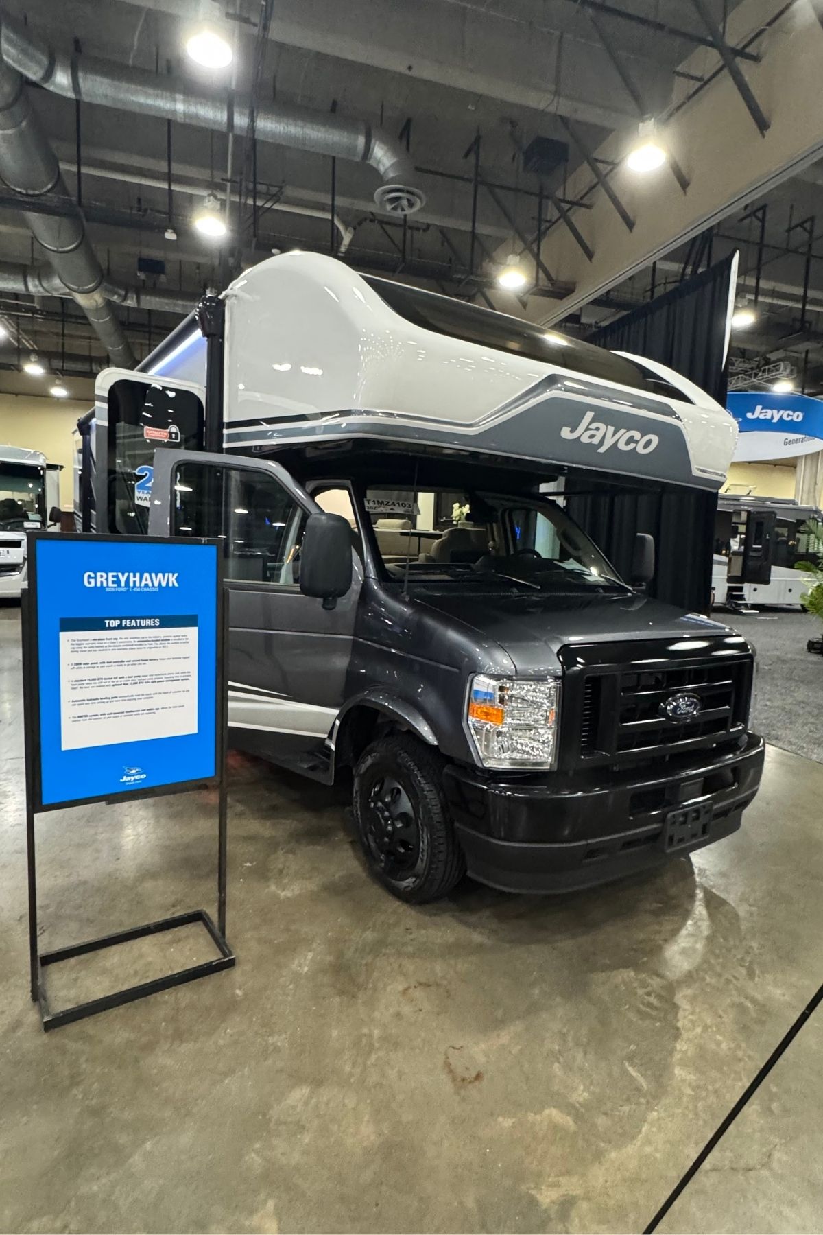 A Jayco Greyhawk, part of the renowned Jayco Class C Motorhomes lineup, is displayed indoors near a blue information sign with specifications.