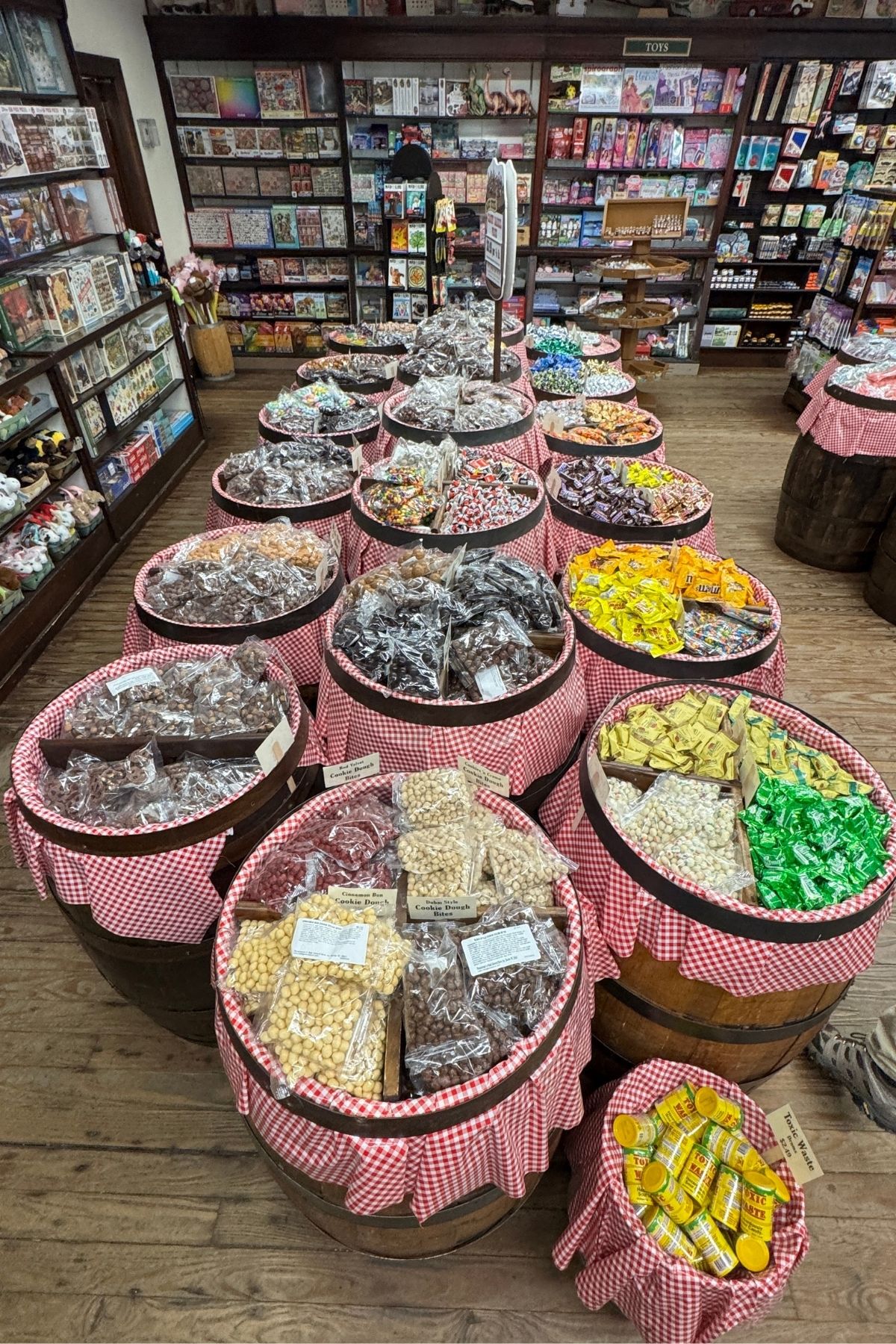 Wooden barrels filled with assorted candies and snacks, some labeled Green Mountain Thousand Trails, are arranged in rows with red gingham cloth covers inside a vintage-style candy store. Shelves lined with packaged goods are in the background.