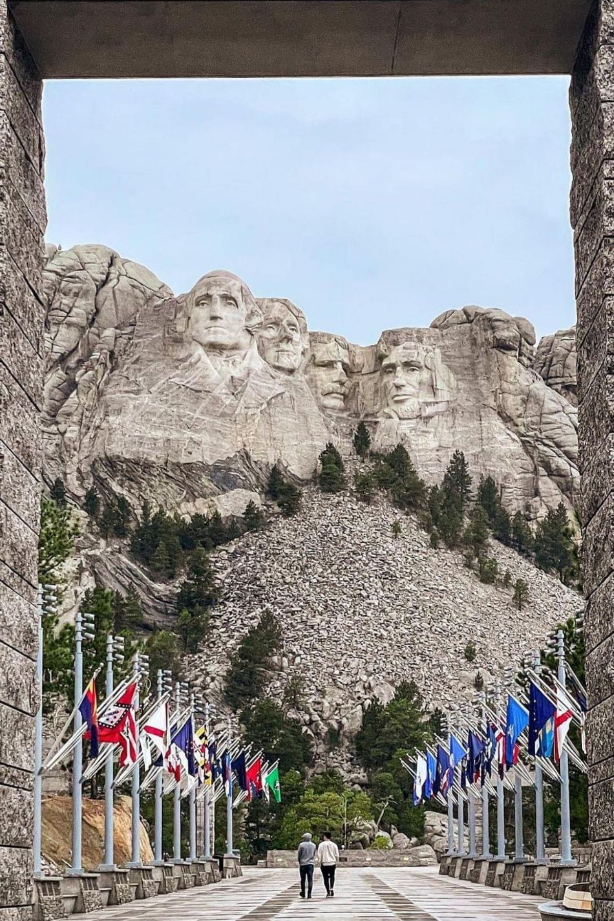 View of Mount Rushmore through a stone archway, with two people walking along a flag-lined pathway toward the monument, capturing the spirit of America as the semiquincentennial approaches.
