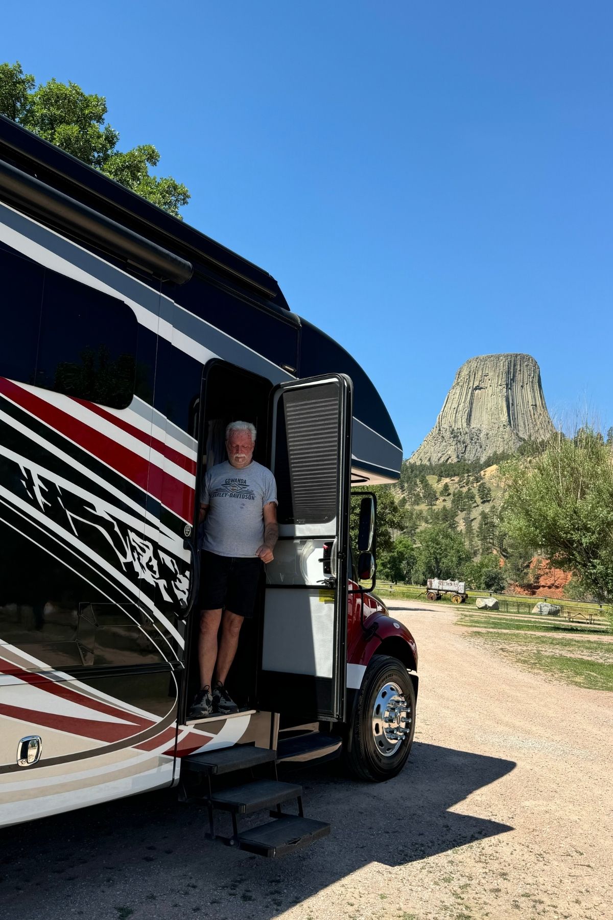 An older man stands in the doorway of a parked RV with striped graphics, celebrating the semiquincentennial, with Devils Tower visible in the background under a clear blue sky.
