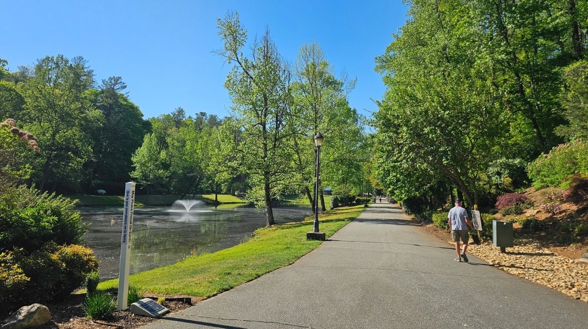 A paved path runs alongside a pond with a fountain, surrounded by trees under a clear blue sky at Green Mountain Thousand Trails; a person walks on the right side of the path.