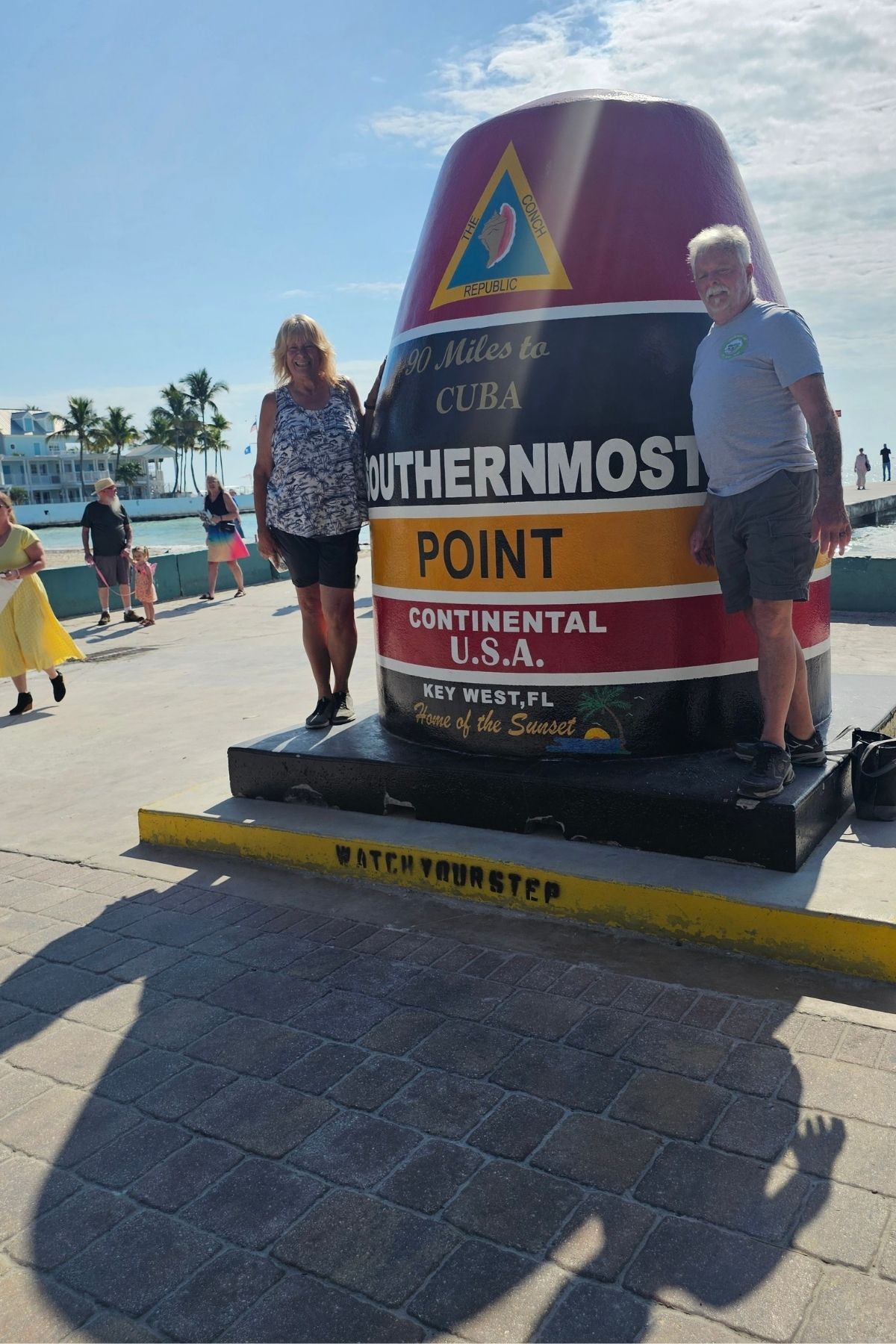 Two people stand next to the Southernmost Point buoy in Key West, a highlight on any Florida Keys RV itinerary, with tourists and palm trees in the background.