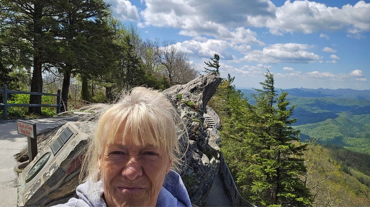 A woman takes a selfie outdoors near a rocky outcrop and trees at Green Mountain Thousand Trails, with distant mountains and a partly cloudy sky in the background.
