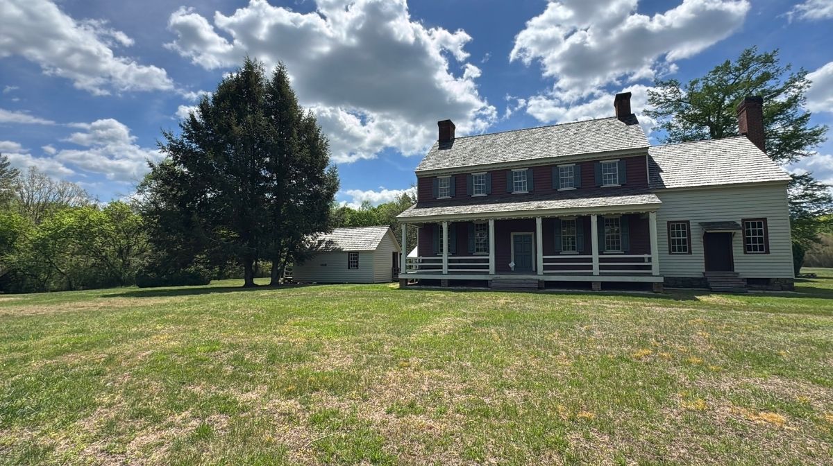 A two-story historic house with a large covered porch sits on a grassy lawn at Green Mountain Thousand Trails, under a partly cloudy sky, with a tree and a smaller building nearby.