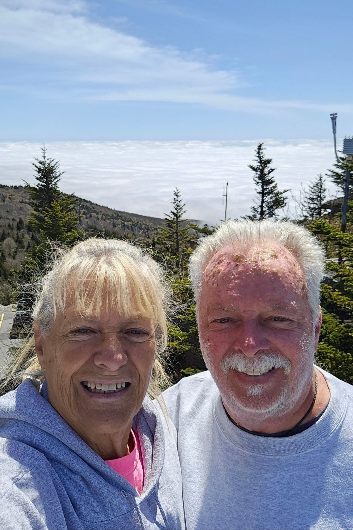 Two older adults smiling for a selfie outdoors at Green Mountain Thousand Trails, with evergreen trees and a cloudy sky in the background.