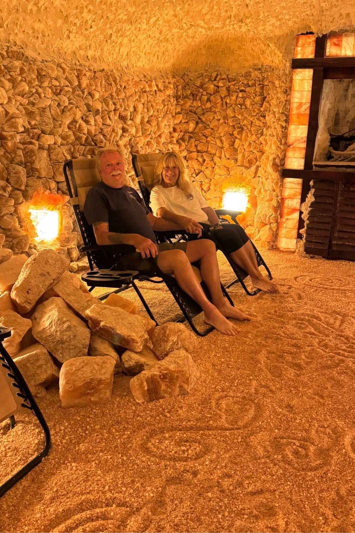Two people relax in reclining chairs inside a salt cave at Green Mountain Thousand Trails, surrounded by salt rocks and warmly lit lamps, with textured salt walls and floor.