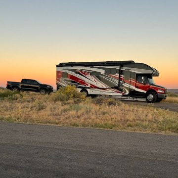 A red and gray RV with black and white designs, perfect for full-time RV living, is parked on a grassy roadside at sunset, with a black pickup truck hitched behind it.