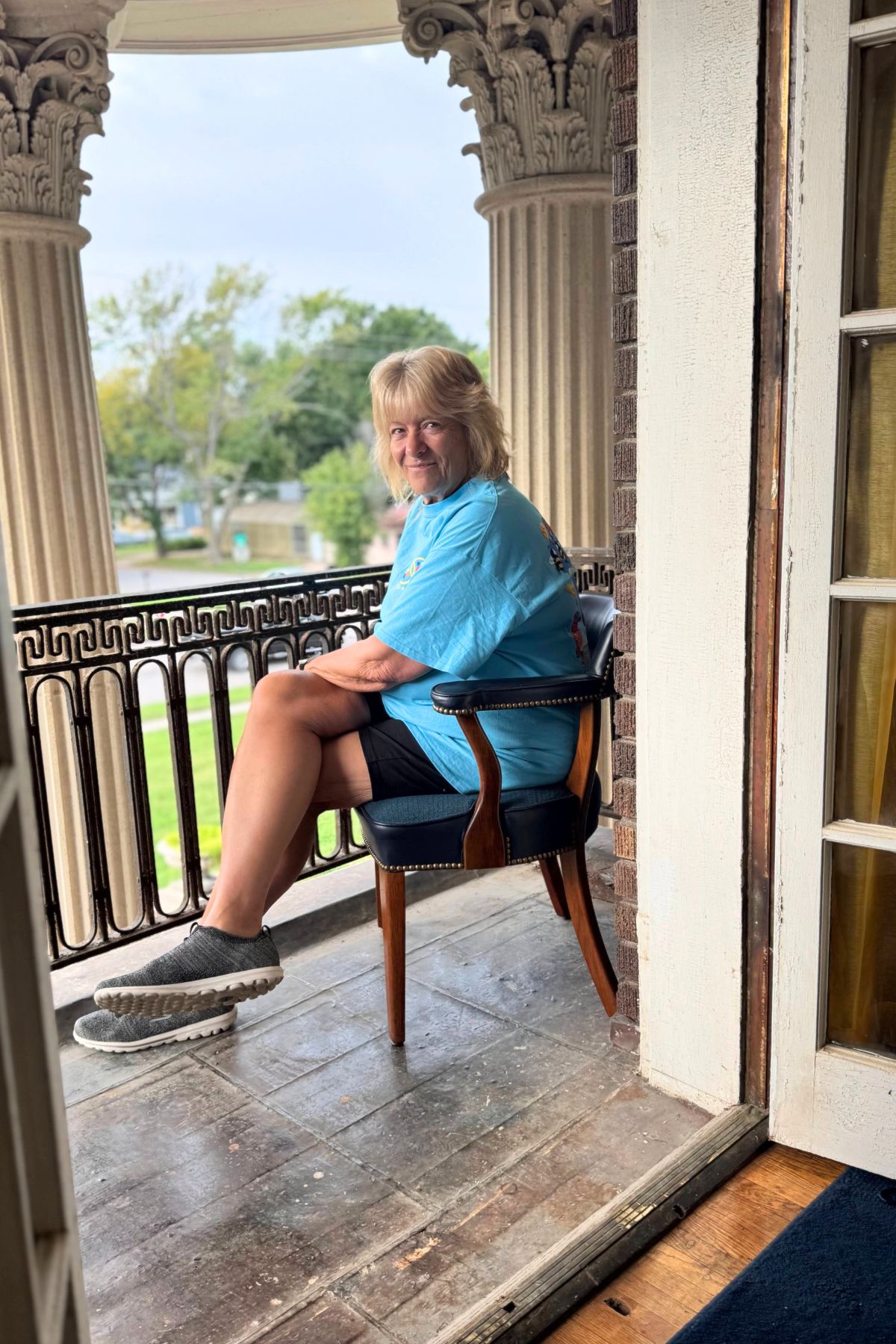 A person wearing a blue shirt and shorts sits on a chair on a balcony with ornate columns, looking toward the camera. Trees and grass stretch into the background, capturing the serene charm of Southeast Kansas.