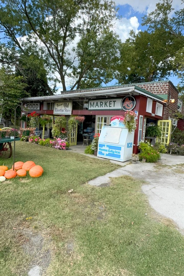 A small market named "Old Riverton Store" with a red exterior, flower displays, pumpkins on the lawn, and an ice freezer by the entrance—nestled amid trees and greenery in the heart of Southeast Kansas.