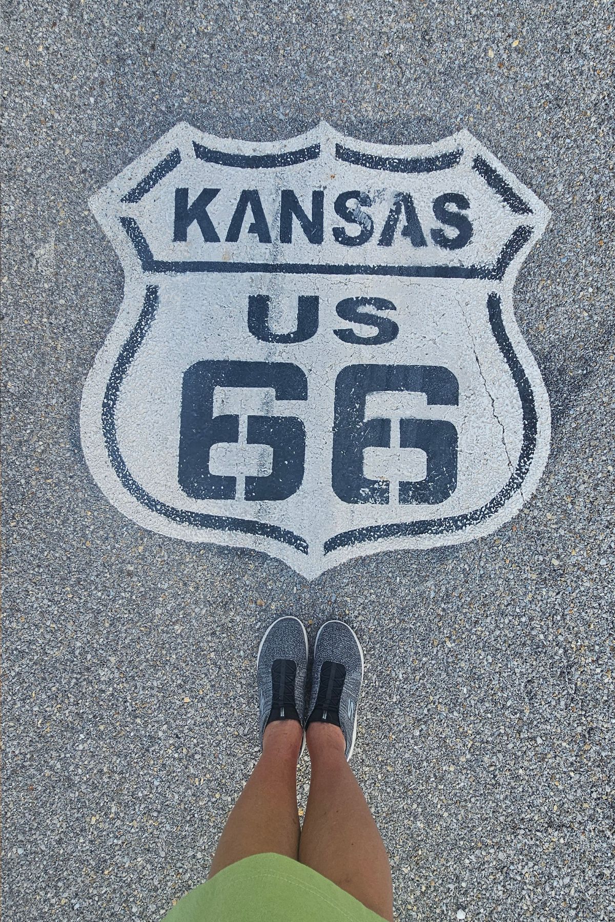 A person stands on the pavement in Southeast Kansas near a painted Route 66 highway emblem with "KANSAS US 66" written inside a shield.