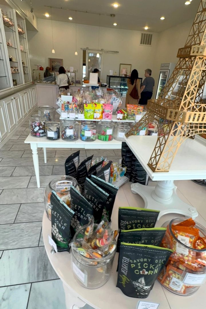 A candy store interior in Southeast Kansas with various jars and bags of sweets on display tables, three people near the counter, and a decorative Eiffel Tower centerpiece.