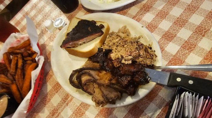 A plate of Southeast Kansas barbecue with brisket, pulled pork, a slice of smoked meat on bread, and a knife sits next to a basket of sweet potato fries on a checkered tablecloth.