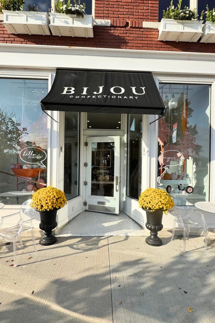 Street view of Bijou Confectionary entrance in Southeast Kansas, featuring a black awning, two large planters with yellow flowers, and clear chairs on either side of the door.
