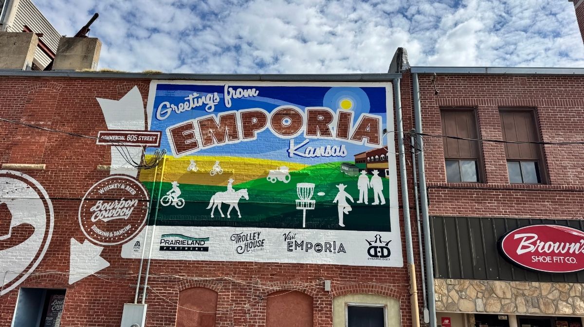 A mural on a brick building in Southeast Kansas reads “Greetings from Emporia, Kansas,” featuring images of local activities and landmarks, including bikes, horses, and a prairie landscape.