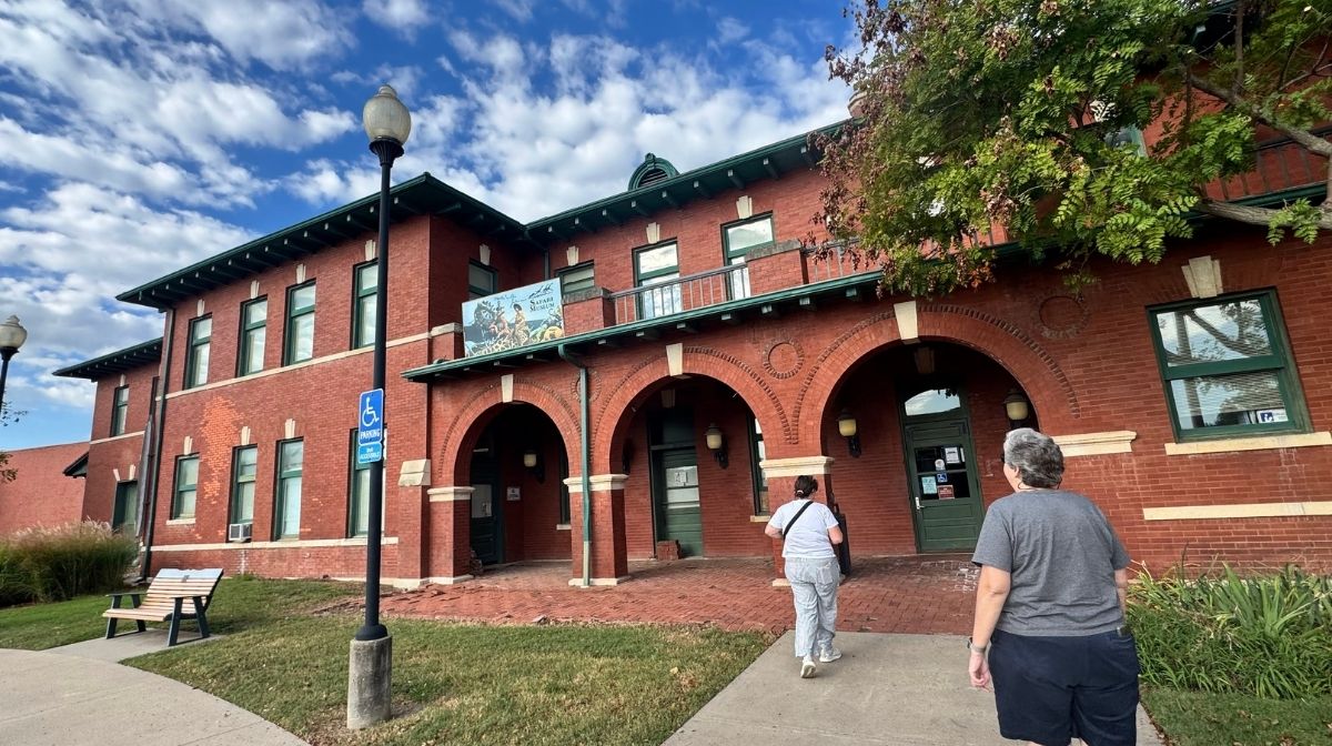 Two people walk toward a red brick building with arched doorways and green trim under a partly cloudy Southeast Kansas sky.