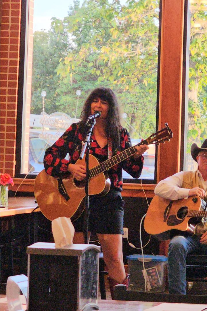 A woman is playing an acoustic guitar and singing into a microphone in front of large windows; beside her, another person with a guitar sits, capturing the relaxed, creative spirit of Southeast Kansas.