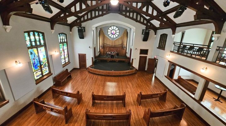 Interior of a Southeast Kansas church with wooden pews, stained glass windows, a stage with musical instruments, and exposed wooden beams on the ceiling.