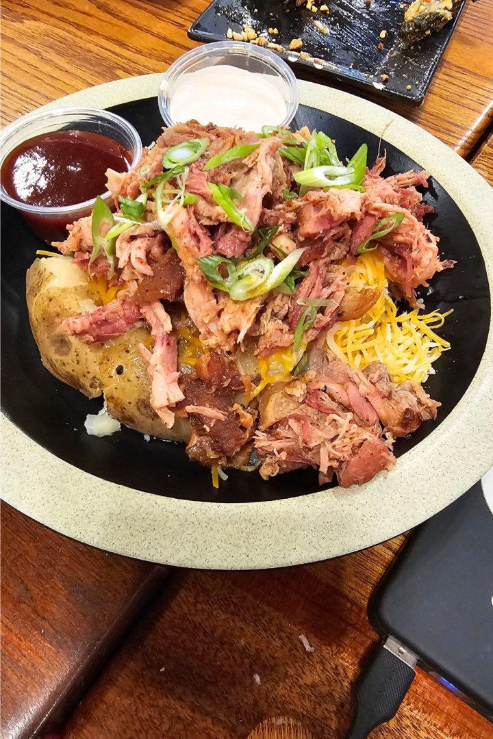 A Southeast Kansas favorite: a baked potato topped with shredded barbecue meat, shredded cheese, and sliced green onions, served with barbecue sauce and a creamy sauce on the side.