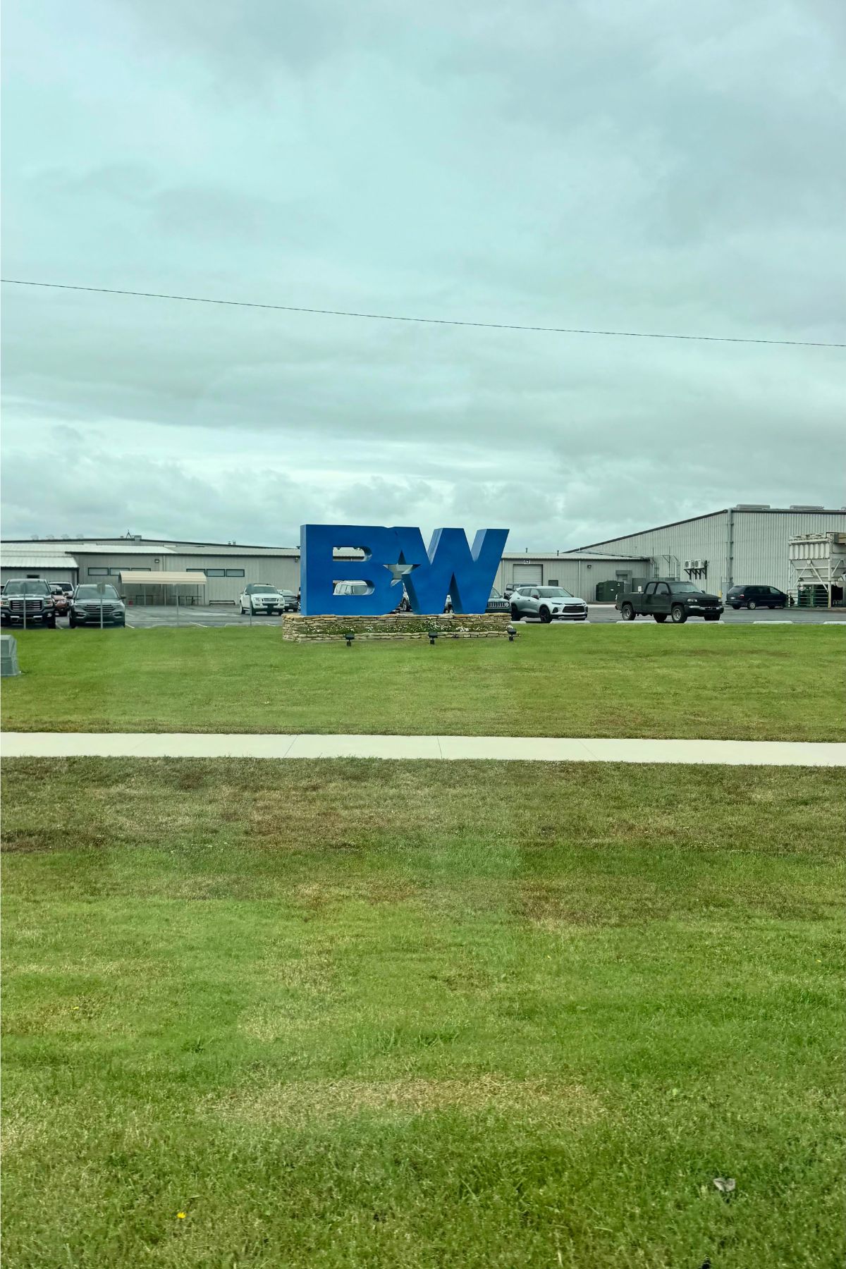 Large blue "BW" sign on a grassy lawn in Southeast Kansas, set before an industrial building with parked cars and a cloudy sky in the background.