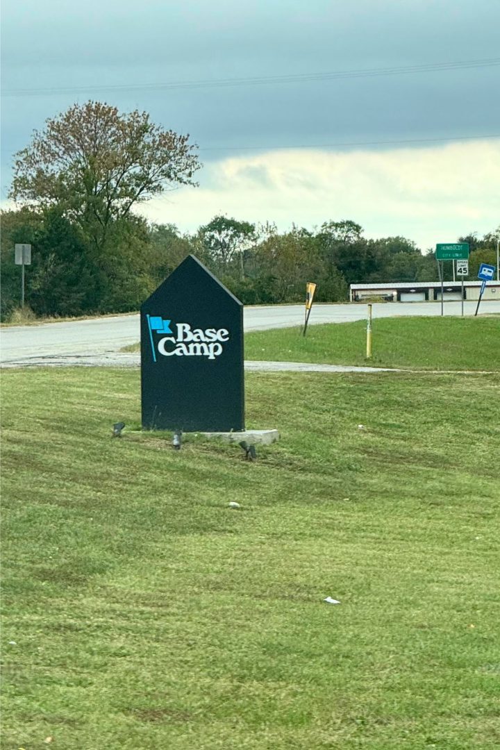 A black sign on a grassy area reads "Base Camp" with a blue flag icon, marking this spot in Southeast Kansas. Trees and road signs are visible in the background under a cloudy sky.