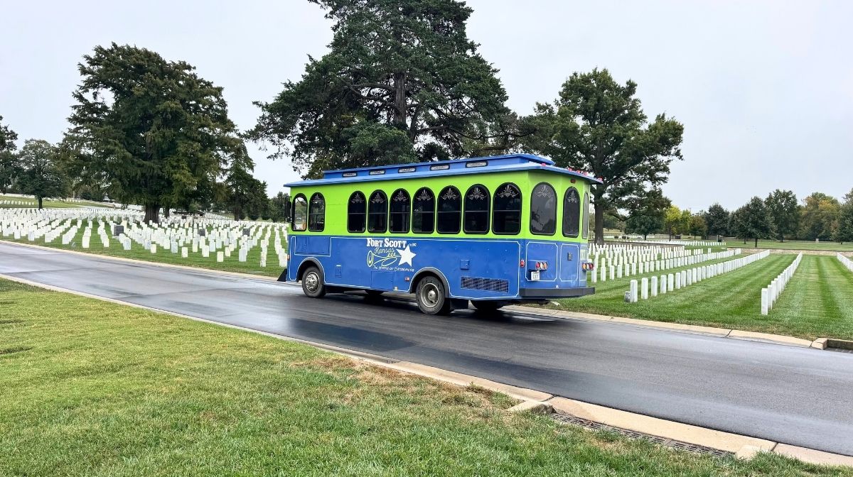 A blue and green tour trolley drives through a Southeast Kansas cemetery with rows of white headstones and green grass on a cloudy day.