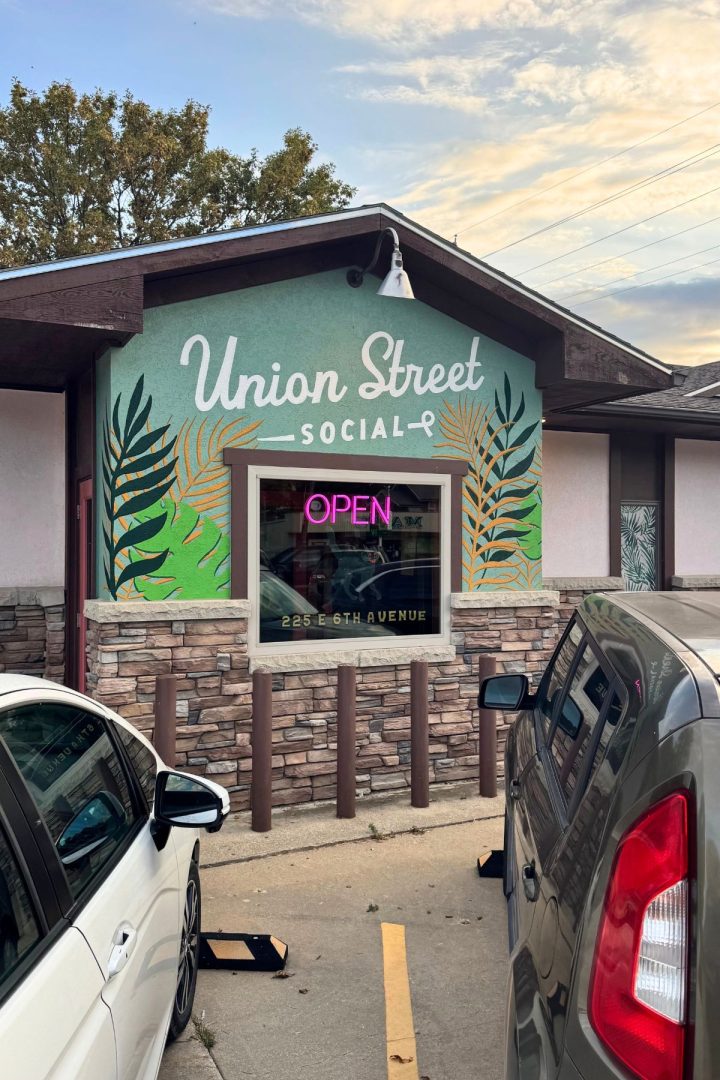 Exterior of Union Street Social in Southeast Kansas, featuring a neon "OPEN" sign, leafy mural, parked cars, and the address 225 E. 6th Avenue visible.