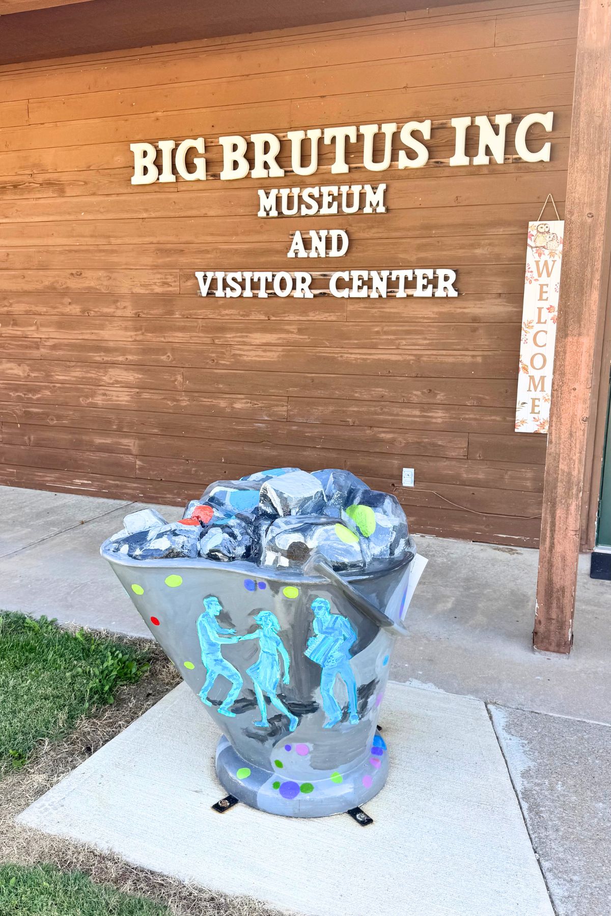 A painted coal cart filled with rocks stands outside the Big Brutus Inc Museum and Visitor Center in Southeast Kansas, next to a wooden welcome sign.