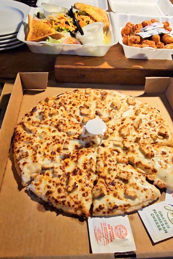 A box of cheesy breadsticks cut into squares with dipping sauces, a takeout container of salad, and another with chicken wings in the background—classic flavors perfect for a Southeast Kansas gathering.