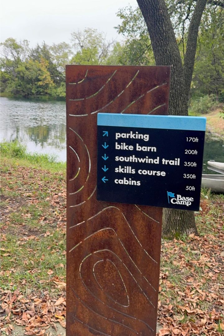A metal sign near a pond in Southeast Kansas lists directions and distances to parking, a bike barn, Southwind Trail, skills course, and cabins at Base Camp.