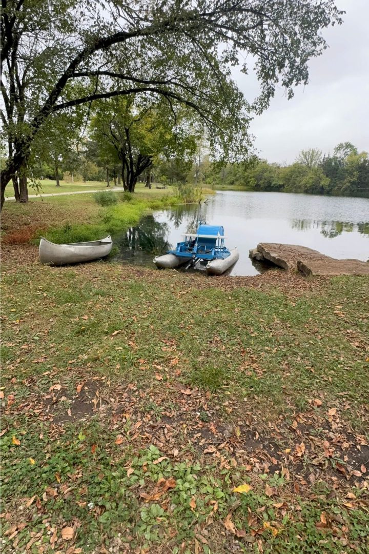 A canoe and a blue pedal boat are positioned at the edge of a calm pond in Southeast Kansas, with trees and grassy areas in the background.