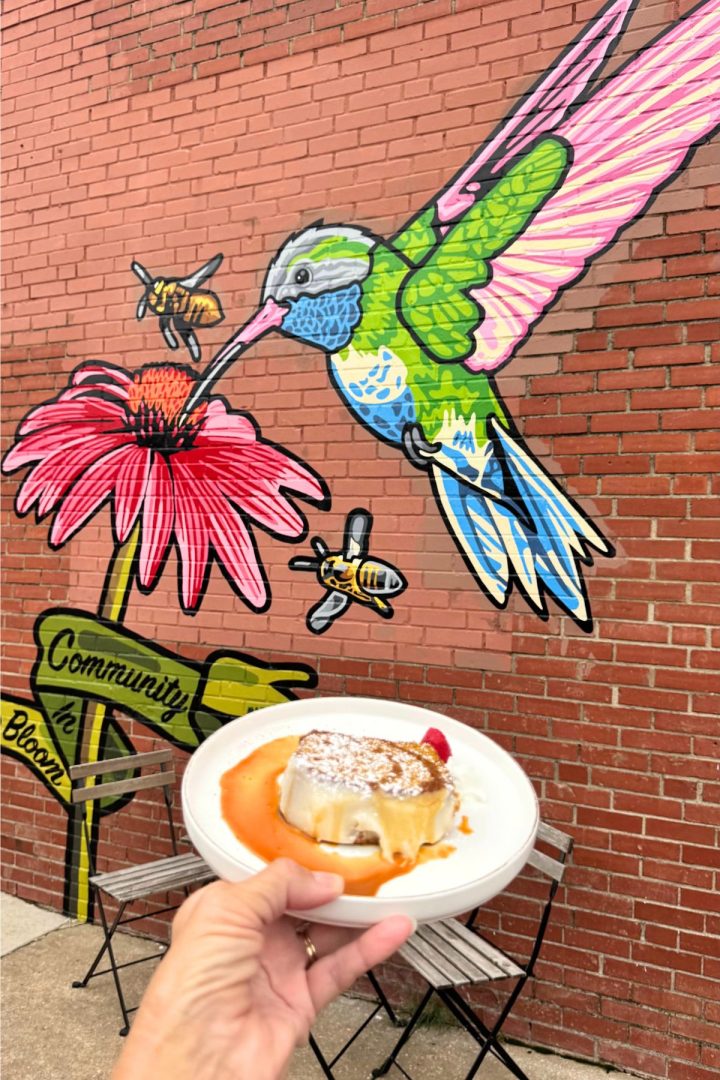 A hand holds a plate with flan and berries in front of a brick wall mural featuring a large hummingbird, bees, and a pink flower—capturing the vibrant charm of Southeast Kansas. Two metal chairs and a table are nearby.