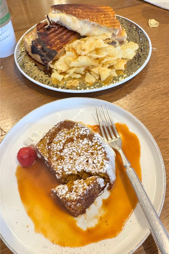 A classic Southeast Kansas breakfast: a plate with a sandwich and potato chips sits behind French toast topped with powdered sugar, a raspberry, and syrup, with a fork resting on the plate.