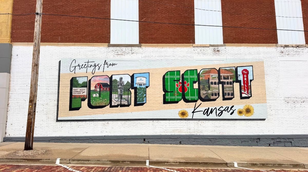 A mural on a white brick wall displays "Greetings from Fort Scott Kansas" with local Southeast Kansas images inside the large letters and sunflowers at the bottom.