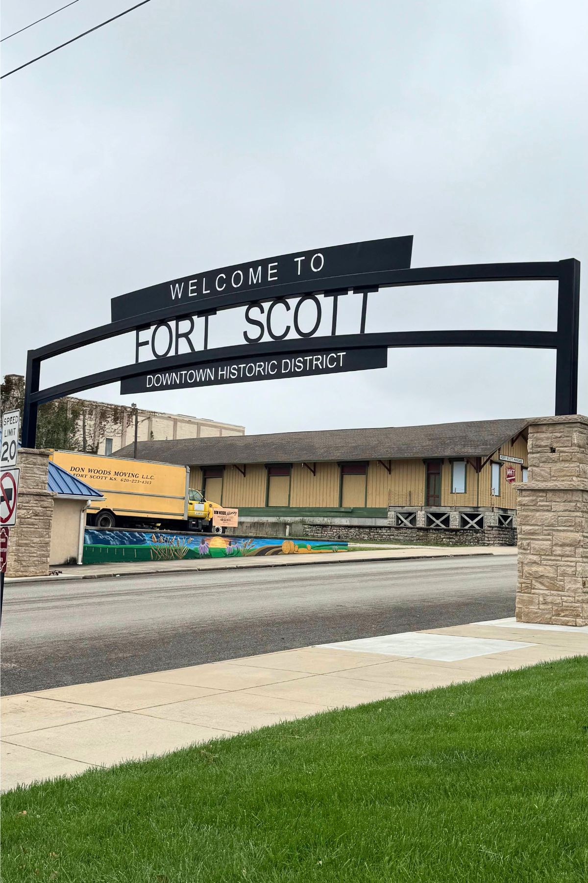 A black metal sign reading "Welcome to Fort Scott Downtown Historic District" arches over a street in Southeast Kansas, with buildings and a truck in the background.