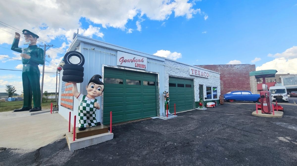 A vintage-style garage in Southeast Kansas with green doors, Texaco signage, large mechanic and Bob’s Big Boy statues, and a classic gas pump in front. Several old cars are parked nearby under a blue sky.