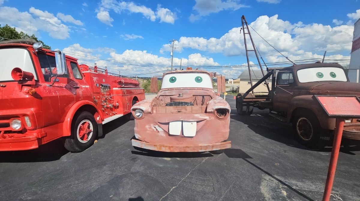 Three old vehicles are parked outdoors in Southeast Kansas; two are tow trucks with cartoon eyes on their windshields, and one is a red fire truck, all under a blue sky with clouds.