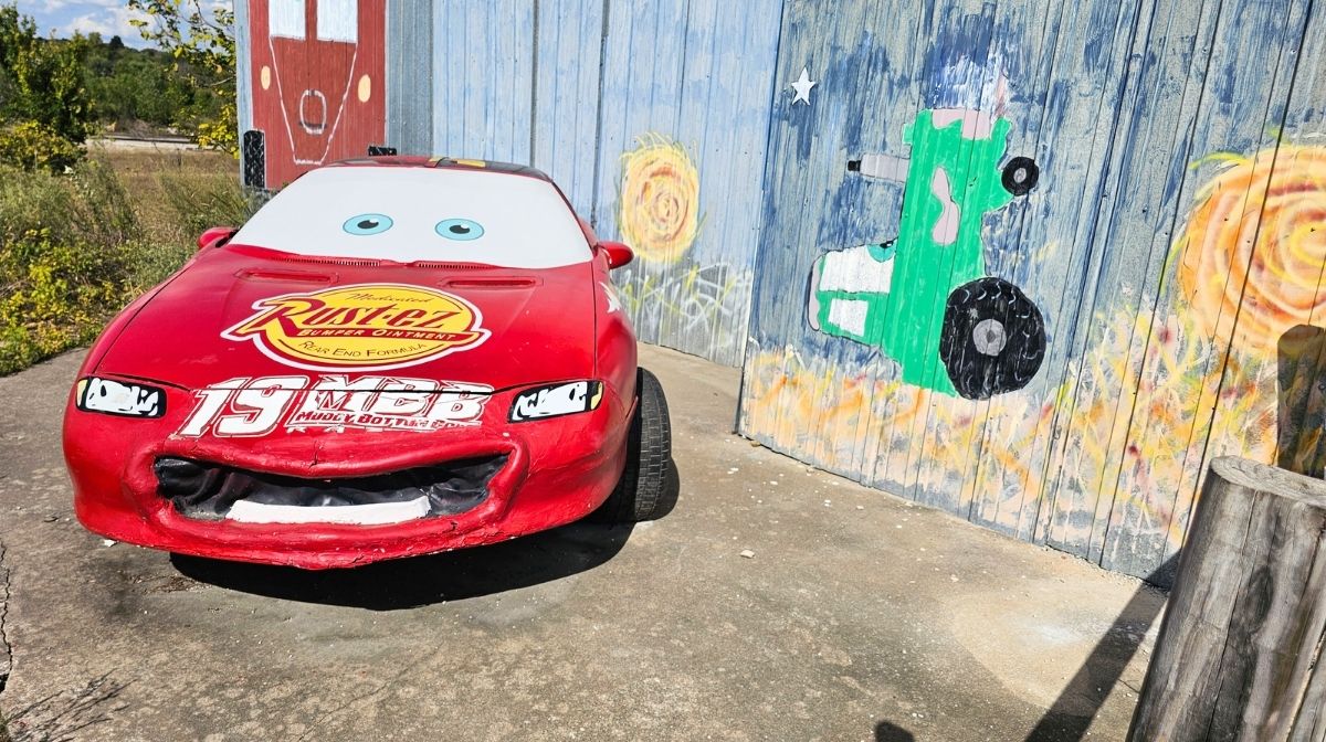 A red cartoon car with large eyes and a "Rust-eze" logo is parked next to a wall in Southeast Kansas, painted with a green tractor and sunflowers.