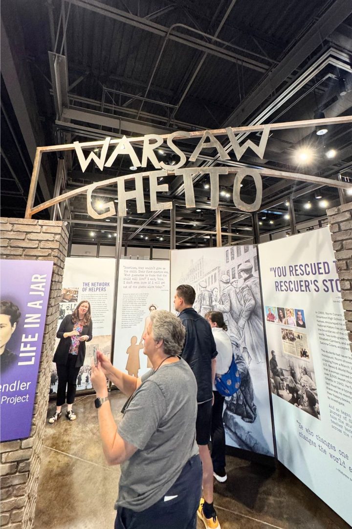 People visit an exhibit about the Warsaw Ghetto in Southeast Kansas, featuring historical panels, photos, and informational displays beneath a large metal sign reading "Warsaw Ghetto.