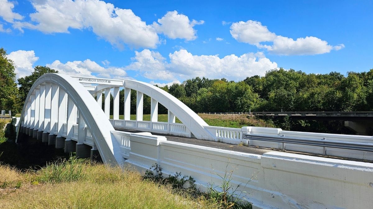 A white arched concrete bridge spans a river in Southeast Kansas, surrounded by green trees under a blue sky with scattered clouds.