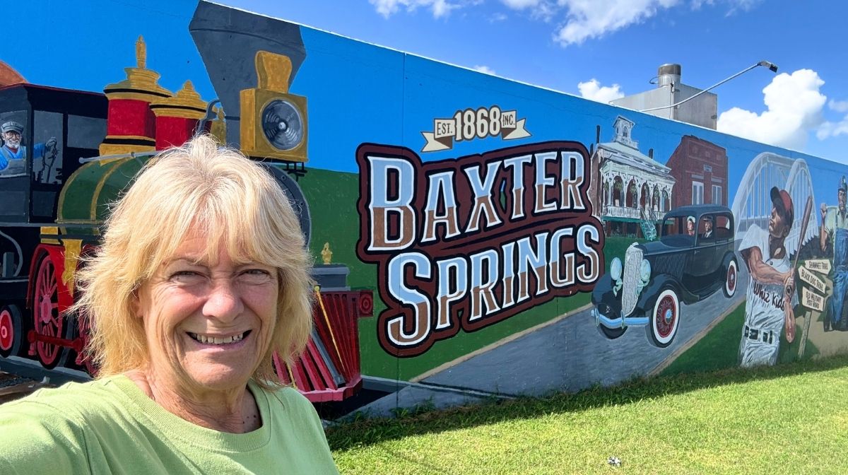 A woman smiles in front of a colorful mural featuring a train, an old car, and the words "Baxter Springs Est. 1868" on a sunny day in Southeast Kansas.